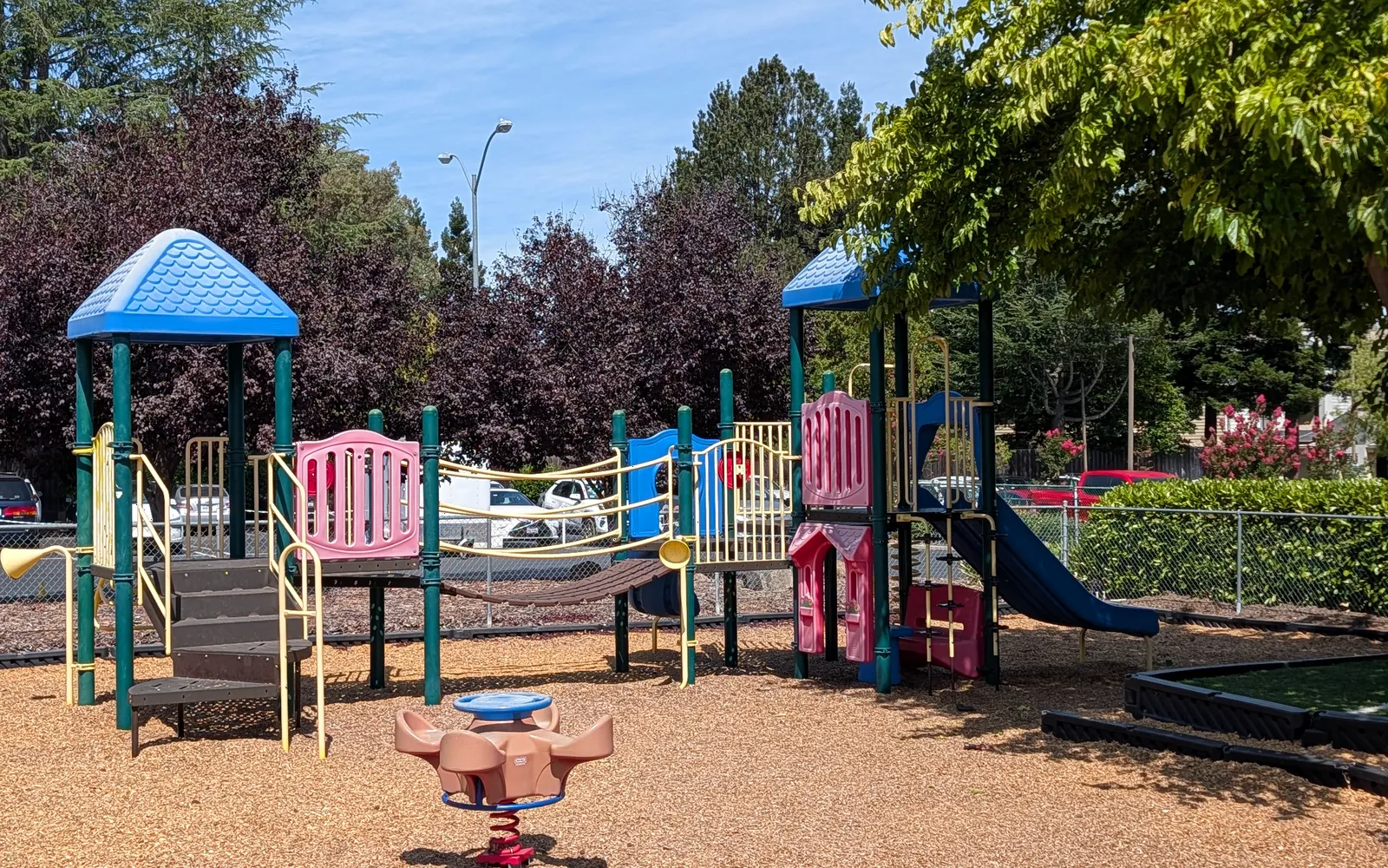 The campus play structure beside the main lot, with a spinner in the bark and shade trees behind.