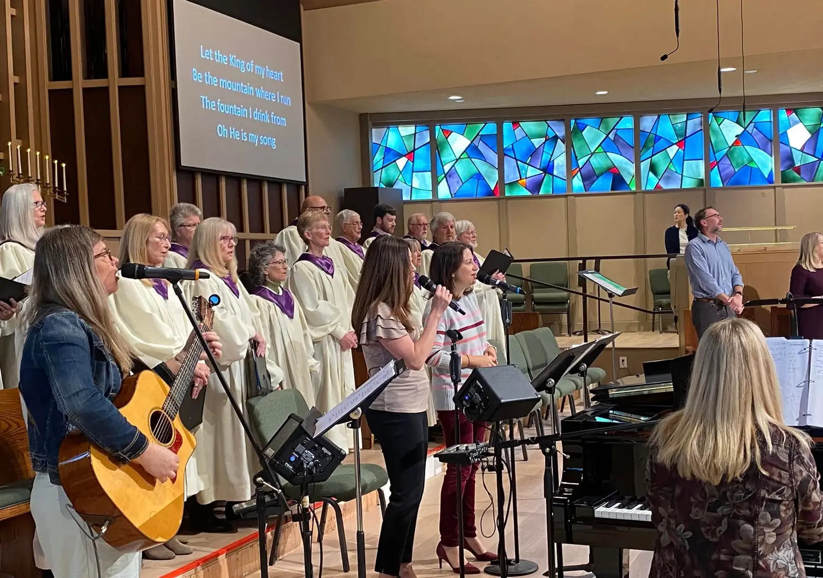 The Chancel Choir in white robes and the worship band with guitar, piano, and singers leading worship in the Sanctuary.