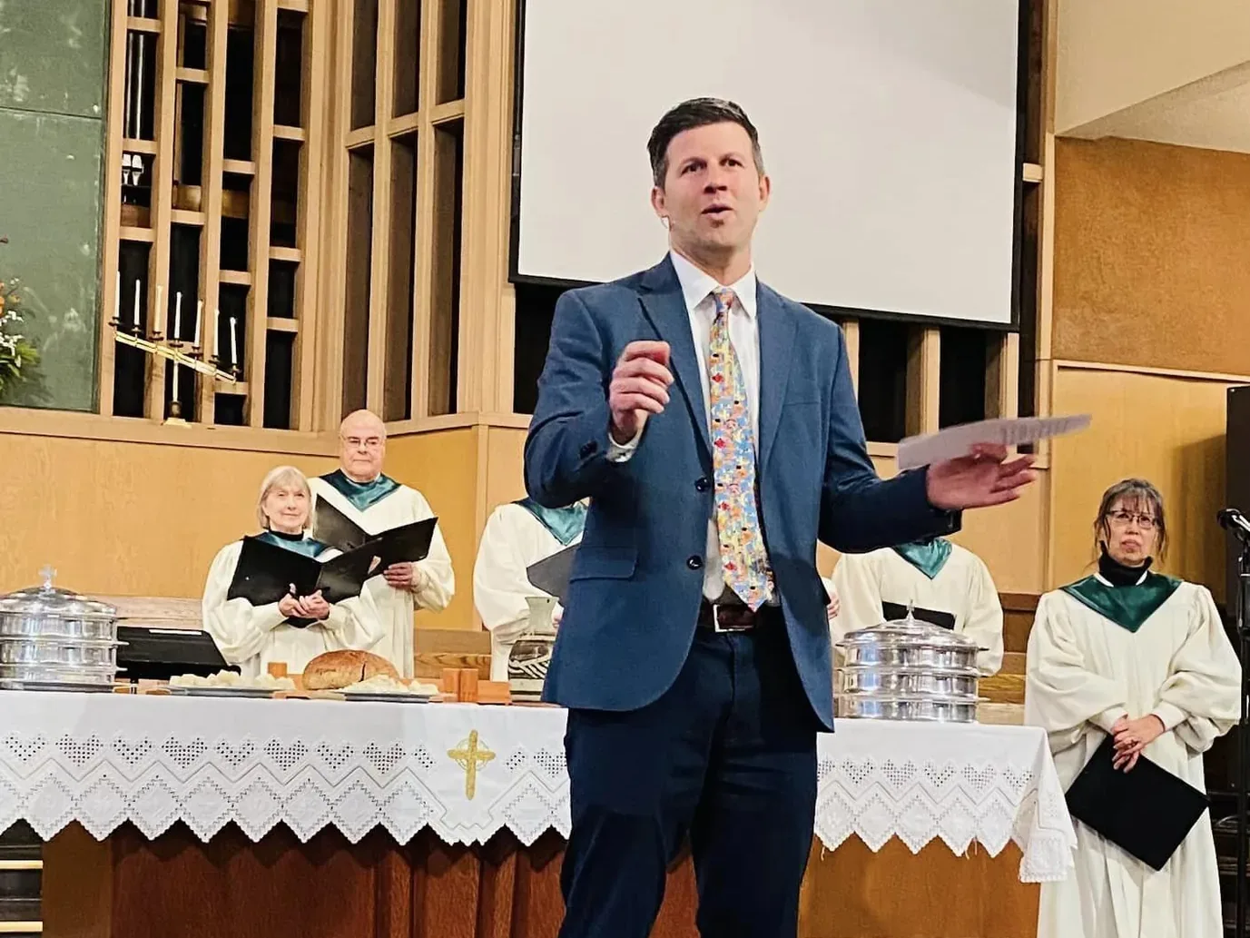 Pastor Dave speaking from beside the communion table, elders in robes holding the elements behind him